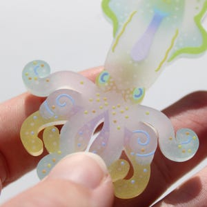 May include: A translucent octopus sticker with pastel colors. The sticker features a light purple body, yellow and blue tentacles, and small yellow and blue dots. The eyes are light blue with a yellow crescent. The tentacles have a swirl design.