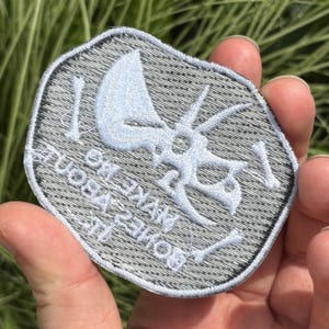 May include: A gray patch with a light blue triceratops skull and bone design. The patch has the text "BONES VIBE MO" in a circular pattern around the design. The patch is held in a hand.
