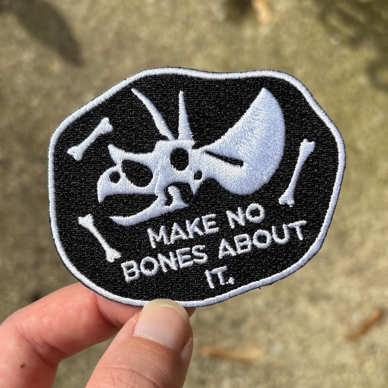 May include: Black and white embroidered patch featuring a Triceratops skull and the phrase "MAKE NO BONES ABOUT IT." The patch has a black border and is likely for clothing or accessories.