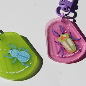May include: Two colorful bug-shaped keychains. One is green with a blue bug, and the other is pink with a multi-colored bug. Both have a purple clasp for attaching to keys or bags.