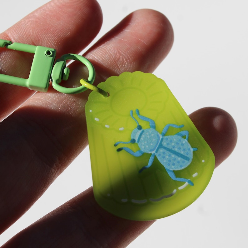 May include: A green keychain with a light blue insect design. The insect has a dotted pattern and is set against a yellow-green background with a sun graphic. The keychain has a green clasp.