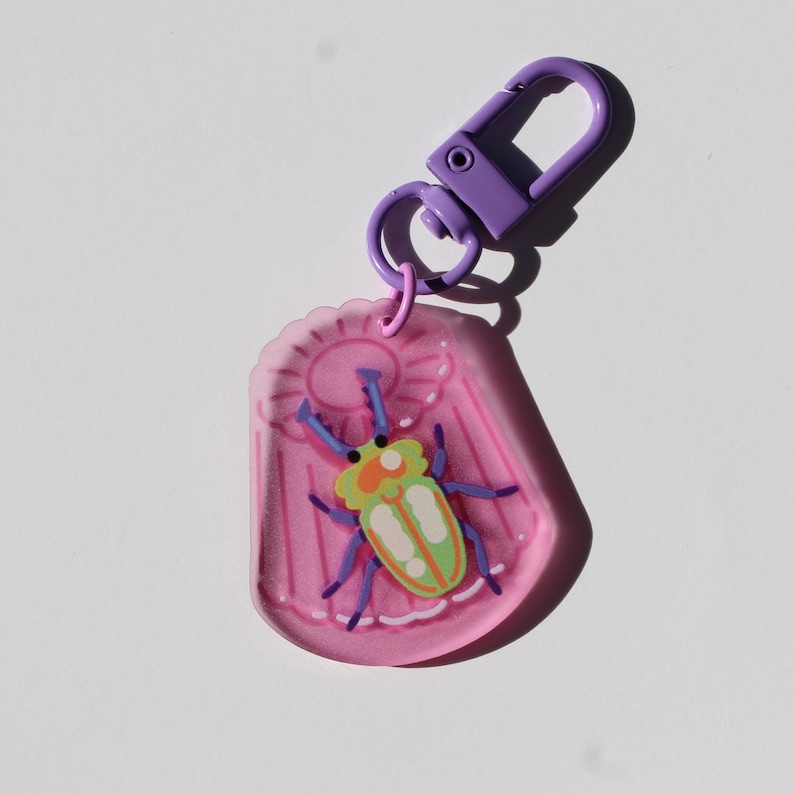 May include: A pink keychain with a purple clasp and ring. The keychain features a cartoon beetle design with green, orange, and blue accents. The beetle is set against a pink background with a sun design.