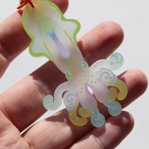 May include: A translucent, colorful squid-shaped charm with a light green outline, yellow and blue accents, and a purple center. The charm is held in a hand and has an orange string attached.