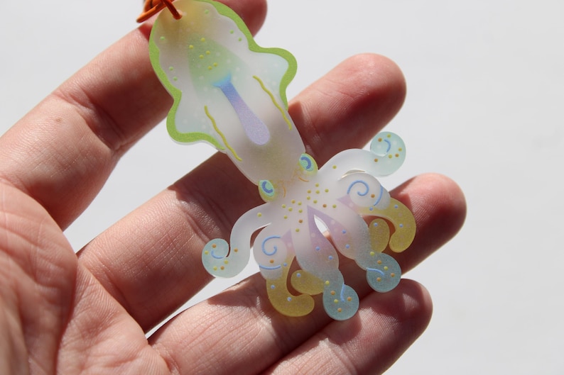 May include: A translucent, colorful squid-shaped charm with a light green outline, yellow and blue accents, and a purple center. The charm is held in a hand and has an orange string attached.