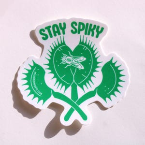 May include: A white sticker with a green Venus flytrap design and the words "STAY SPIKY" in green. The flytrap has a fly in the center. The website address "microbiomearts.com" is printed on the left side of the sticker.