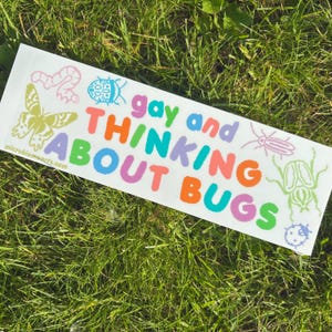 May include: A white rectangular sticker with the text "gay and THINKING ABOUT BUGS" in colorful letters. The sticker features illustrations of various insects and a butterfly. The website address "microbiomearts.com" is printed at the bottom.
