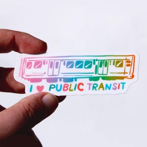 May include: A colorful sticker featuring a stylized subway train illustration. The train is outlined in a rainbow gradient, with the words "I ❤️ PUBLIC TRANSIT" in blue, red, and green. The sticker is held against a white background.