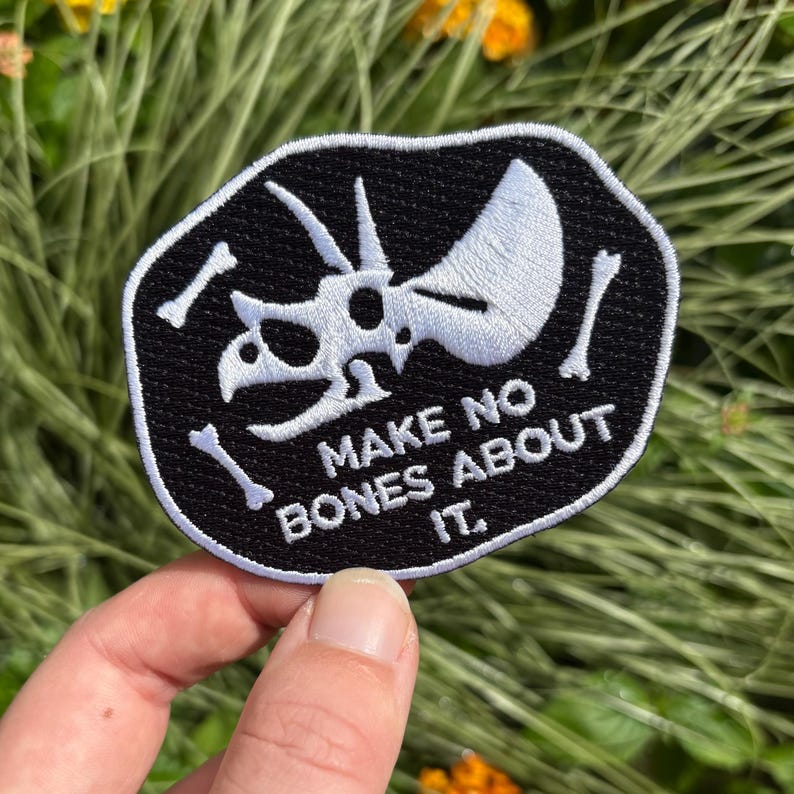 May include: Black patch with a white Triceratops skull and bones design. The text "MAKE NO BONES ABOUT IT" is embroidered in white. The patch is held in a hand against a background of green and yellow foliage.