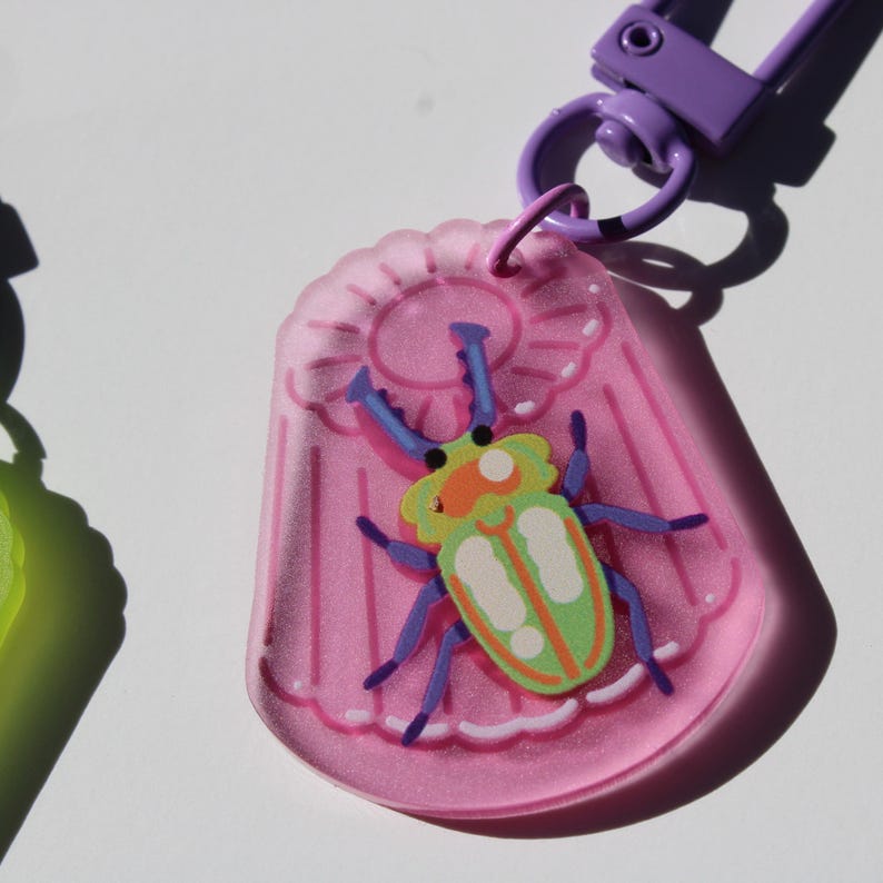 May include: A pink, bug-shaped keychain with a purple clasp. The keychain features a colorful cartoon beetle with green, orange, and blue accents. The background is a plain white surface.