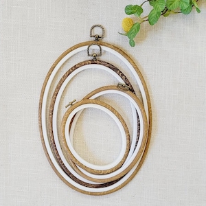 May include: Three wooden embroidery hoops with white inner rings. The hoops are nested together, with the largest hoop at the bottom and the smallest hoop at the top. The hoops are made of natural wood with a brown finish.