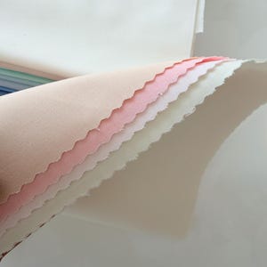 May include: A stack of fabric swatches in various pastel colors, including light pink, white, and peach. The edges of the fabric are scalloped, and the swatches are layered on top of each other. The top swatch is white, and the bottom swatch is a light peach color.