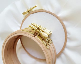May include: Close-up of two wooden embroidery hoops with brass hardware. One hoop is in focus, showing the adjustable screw mechanism. The other hoop is in the background, resting on white fabric.