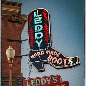 May include: A neon sign for a boot and saddle shop called "Leddy's Boots & Saddlery". The sign is shaped like a boot and has the words "Hand Made Boots" on it. The sign is lit up in red, blue, and green neon lights. The shop is located at N. Main Exchange.