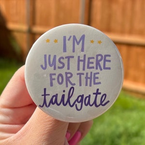 Puede incluir: Botón blanco con texto morado que dice "Solo estoy aquí para el tailgate" con tres estrellas doradas a cada lado del texto.