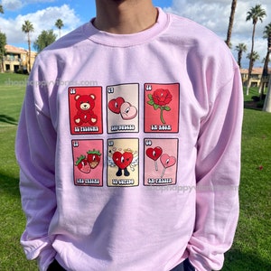 May include: A pink sweatshirt with a graphic print of a Loteria card set. The cards feature illustrations of a teddy bear, candy, a rose, strawberries, Cupid, and a lollipop. The text on the cards reads "El Peluche", "Los Dulces", "La Rosa", "Las Fresas", "El Cupido", and "La Paleta".