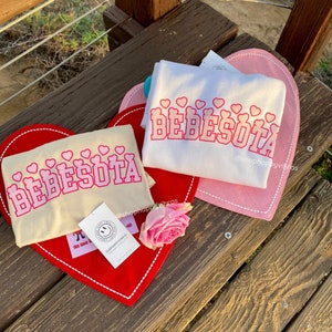 May include: Two beige and white t-shirts with the text "Bebesota" in pink letters and pink hearts. The shirts are displayed on red and pink heart-shaped surfaces. The text "@shophappyvibros" is on both shirts.