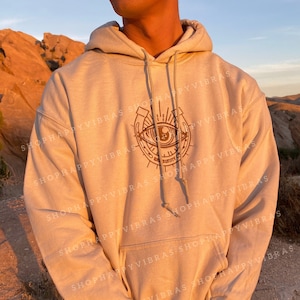 May include: A beige hoodie with a brown graphic of an eye inside a horseshoe with the text "Shop Happy Vibes" repeated around the graphic.