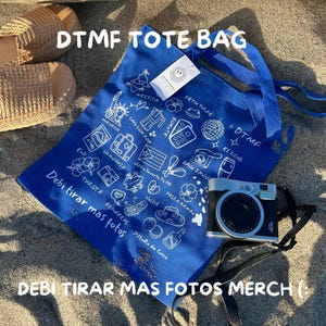 May include: A blue tote bag with white illustrations and text, including "DTMF TOTE BAG" and "Debi tirar mas fotos merch (:)". A black and silver camera rests beside the bag on a sandy surface. Woven sandals are visible in the background.