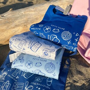 K&ouml;nnte beinhalten: Ein Stapel blauer und hellblauer T-Shirts und eine Tragetasche mit wei&szlig;en grafischen Designs. Die Designs umfassen eine Discokugel, Blumen und Text. Die Tragetasche hat Text auf Spanisch. Ein rosa Sweatshirt ist im Hintergrund zu sehen.