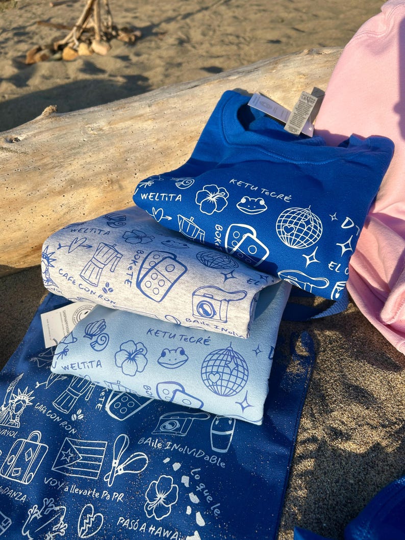 K&ouml;nnte beinhalten: Ein Stapel blauer und hellblauer T-Shirts und eine Tragetasche mit wei&szlig;en grafischen Designs. Die Designs umfassen eine Discokugel, Blumen und Text. Die Tragetasche hat Text auf Spanisch. Ein rosa Sweatshirt ist im Hintergrund zu sehen.