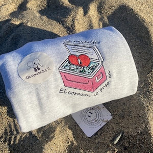 May include: A gray sweatshirt with a pink cooler and a red heart inside. The text "La neverita" is above the cooler and "El corazon lo puso en" is below the cooler. A white circle with a cartoon figure and the text "On Verano Sin Ti" is on the left side of the sweatshirt. A white square with a smiley face and the text "Shop Happy Vibras" is in the bottom right corner of the image.