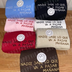 May include: Six different colored sweatshirts with the same text embroidered on the front. The text reads "Nadie sabe lo que va a pasar mañana". Each sweatshirt has a small smiley face logo in the top left corner.