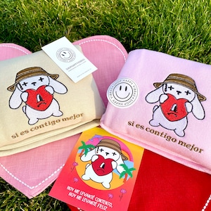 May include: Two beige and pink fabric pouches with embroidered white bunny holding a red heart. The pouches have the text "si es contigo mejor" on them. A red fabric pouch with a white bunny holding a red heart and the text "HOY ME LEVANTÉ CONTENTO, HOY ME LEVANTÉ FELIZ" is in front of the pouches.