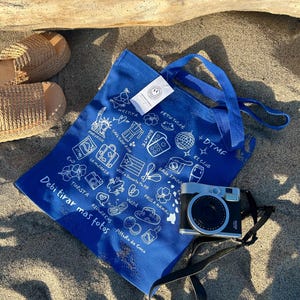 K&ouml;nnte beinhalten: Eine blaue Tragetasche mit wei&szlig;en Illustrationen und Text, darunter "Debi tirar mas fotos." Eine silberne und schwarze Kamera mit schwarzem Riemen liegt auf dem Sand. Geflochtene Espadrilles sind ebenfalls zu sehen.