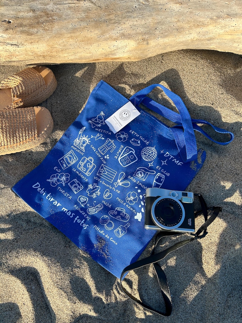 K&ouml;nnte beinhalten: Eine blaue Tragetasche mit wei&szlig;en Illustrationen und Text, darunter "Debi tirar mas fotos." Eine silberne und schwarze Kamera mit schwarzem Riemen liegt auf dem Sand. Geflochtene Espadrilles sind ebenfalls zu sehen.