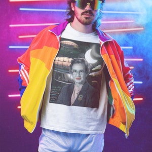 May include: A person wearing a white t-shirt with a graphic of a woman and airships. The t-shirt has the text "Katherine Johnson,  NASA,  "We calculate the stars,  we walk on the moon,  we balance the books,  except for the books,  we don't do that."  The person is also wearing a rainbow colored jacket and white athletic shorts.