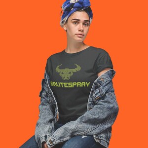 May include: A person wearing a black t-shirt with a green pixelated bull head and the text "BRUTESPRAY" on it. They are wearing a blue denim jacket and blue jeans.