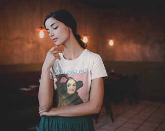 May include: A white t-shirt with a colorful graphic of Ada Lovelace, a pioneer in computer programming. The graphic includes a portrait of Ada Lovelace with a pink flower in her hair. The text "Ada Lovelace" is printed in blue and pink letters on the shirt.