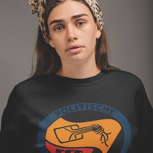 May include: A black sweatshirt with a circular graphic featuring a stylized fist holding a wrench. The graphic is in orange, red, blue, and white. The text "POLITISCHE AKTION" is written around the graphic.
