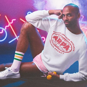 Peut inclure: Une personne portant un sweat-shirt blanc avec le texte "Defense Advanced Research Projects Agency" et un logo rouge avec le texte "ARPA" dans un cercle. La personne porte un short rose et des chaussettes blanches avec des rayures vertes et jaunes.