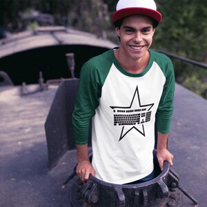 May include: A white and green baseball tee shirt with a black star graphic featuring a keyboard inside the star. The person wearing the shirt is standing in a doorway.