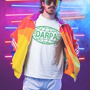 May include: A person wearing a white t-shirt with a green globe graphic and the text "DARPA" inside the globe. The person is wearing a colorful jacket with yellow, orange, red, and blue panels. The person is also wearing white athletic shorts with a black trim.