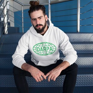 Peut inclure: Un sweat-shirt blanc avec un graphique de globe vert et le texte "DARPA" sur le devant. La personne portant le sweat-shirt est assise sur un escalier en métal.