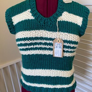 May include: A hand-knitted green and white striped vest. The sleeveless top has a crew neck and is displayed on a mannequin. A tag with text is attached to the vest. The vest is made of a chunky knit yarn.