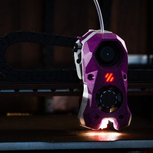 May include: A close-up of a purple 3D printer hot end with a fan and a red LED light. The hot end is attached to a black cable and is sitting on a metal surface.