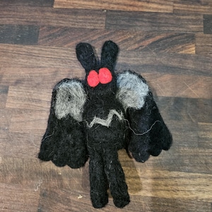 Può includere: Un pipistrello di feltro nero con occhi rossi e ali grigie. Il pipistrello è in piedi su una superficie di legno.