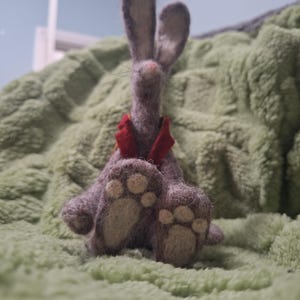 Könnte beinhalten: Ein handgefertigtes Hasenkaninchen aus Filz mit roter Fliege. Das Kaninchen ist grau mit hellbeige Pfotenpolstern. Das Kaninchen sitzt auf einer grünen, strukturierten Oberfläche.