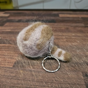 May include: A brown and pink felted cat keyring with a silver key ring. The cat is curled up in a ball with its tail wrapped around its body.