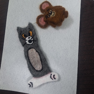 May include: Handmade felted Tom and Jerry characters. The gray Tom cat has a white face and belly, with orange and black details. The brown Jerry mouse has pink ears. Both are connected by a silver chain.