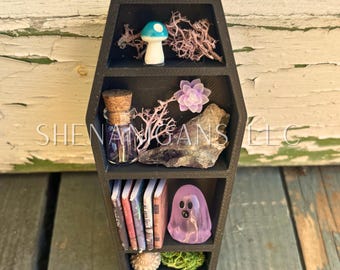 May include: Black coffin-shaped shelf with three compartments, filled with miniature items. Includes a mushroom, books, a ghost figurine, a bottle, and other decorative elements. The text "SHENANIGANS LLC" is visible.