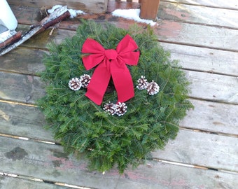 May include: A circular evergreen wreath, adorned with a bright red bow and several pine cones with white tips. The wreath is displayed on a weathered wooden surface, ideal for festive decoration.