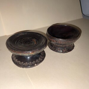 May include: Two handcrafted, dark-colored ceramic bowls with a pedestal base. The bowls have a circular design on the top and decorative patterns on the base. The bowls are likely for decorative purposes.