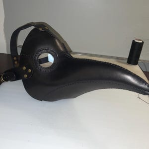 May include: A black leather plague doctor mask with a long, curved beak and eye holes. The mask has a strap for securing it to the head and is adorned with brass rivets. A spool of black thread sits nearby.