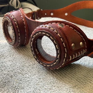 May include: Brown leather steampunk goggles with white stitching and brass accents. The goggles have clear lenses and a brown leather strap. The design features a classic, vintage aesthetic.