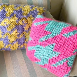 May include: Two knitted throw pillows with houndstooth patterns. One pillow features yellow and purple, the other pink and turquoise. The pillows are on a striped chair, showcasing the handmade texture and vibrant colors.