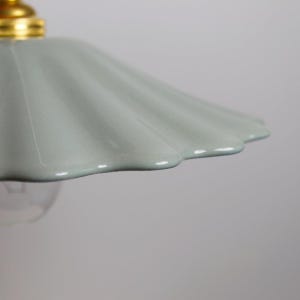 Op de afbeelding: Een close-up van een lichtblauwe keramische lampenkap met een geschulpte rand. De lampenkap heeft een glanzende afwerking en is bevestigd aan een goudkleurige fitting. De gloeilamp is eronder zichtbaar.